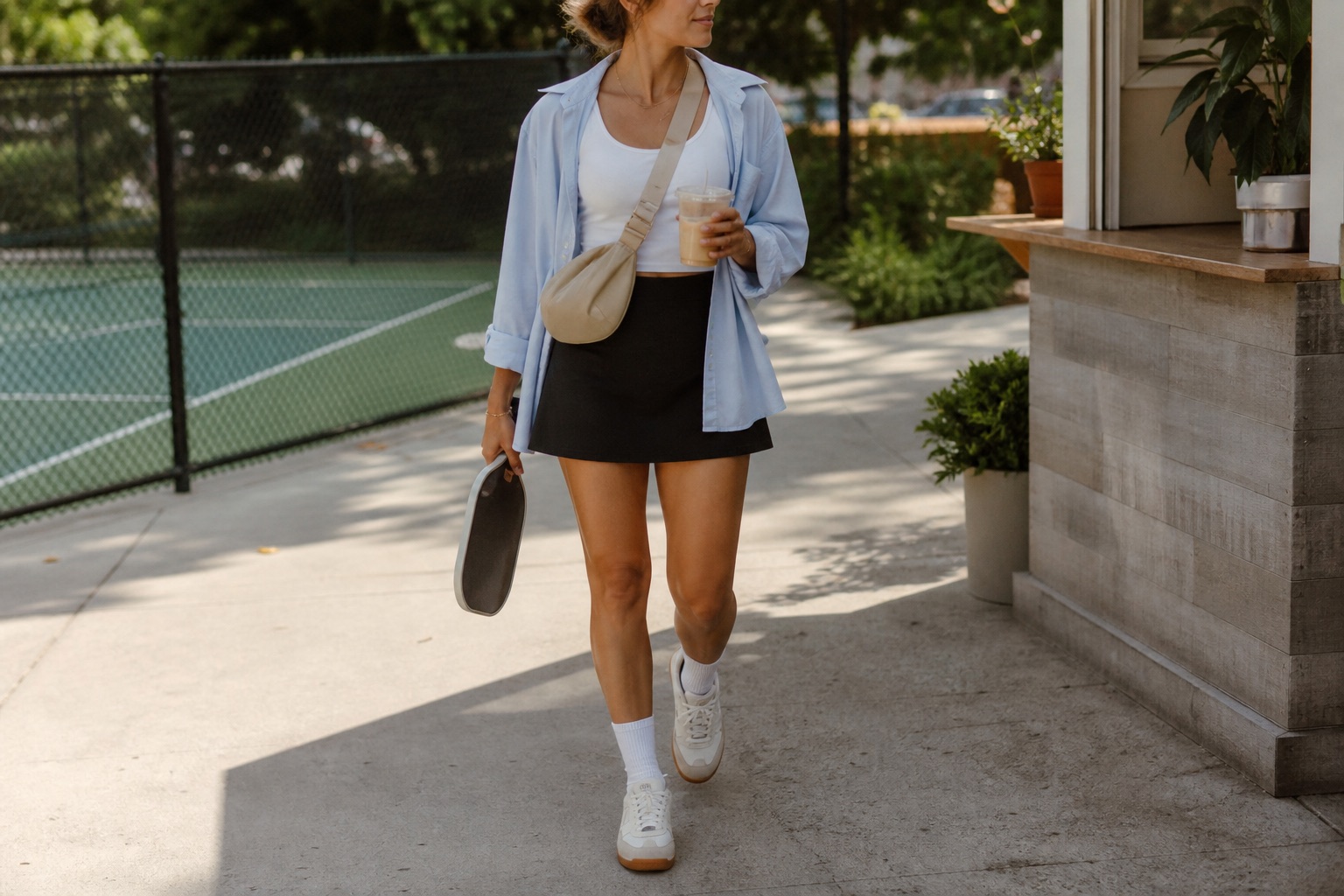 Court-to-coffee pickleball outfit with black skort, white top, pale blue button-down shirt, sneakers, crossbody bag, paddle, and iced coffee